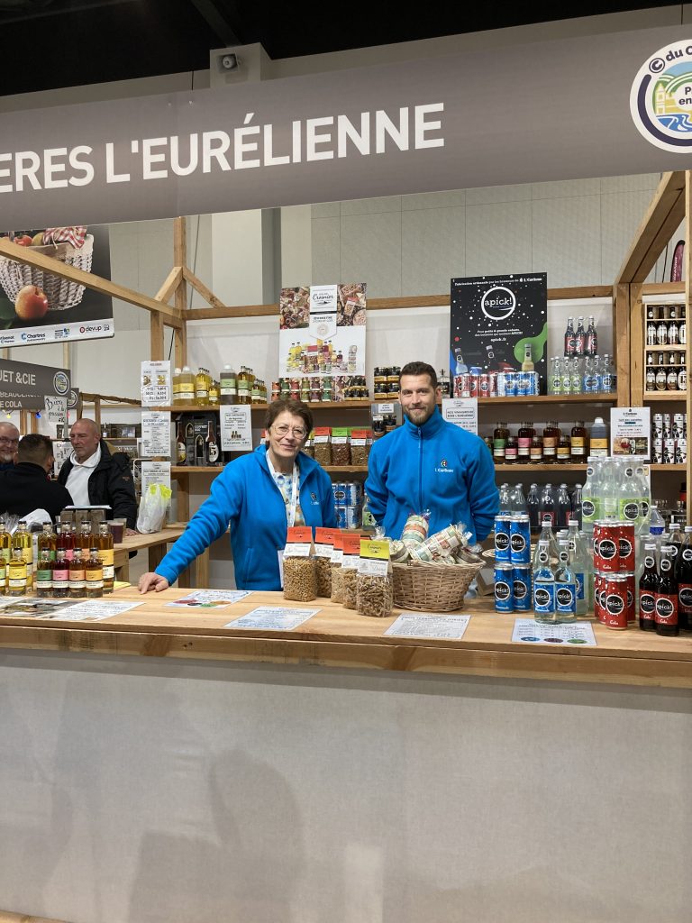 Le stand de l'Eurélienne avec Blandine Crosnier et son fils situés derrière un comptoir sur lequel sont disposés quelques produits de l'Atelier Crosnier. Au fond du stand, des étagères sont remplis de bouteilles de bières, pâtes, huiles et autres produits de leur fabrication.