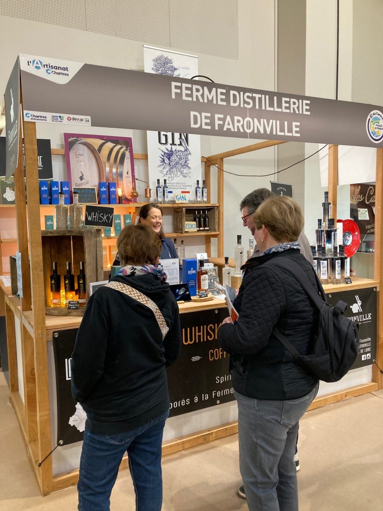 Stand de la distillerie de Faronville avec des étagères bois au fond garnies de bouteilles de gin.