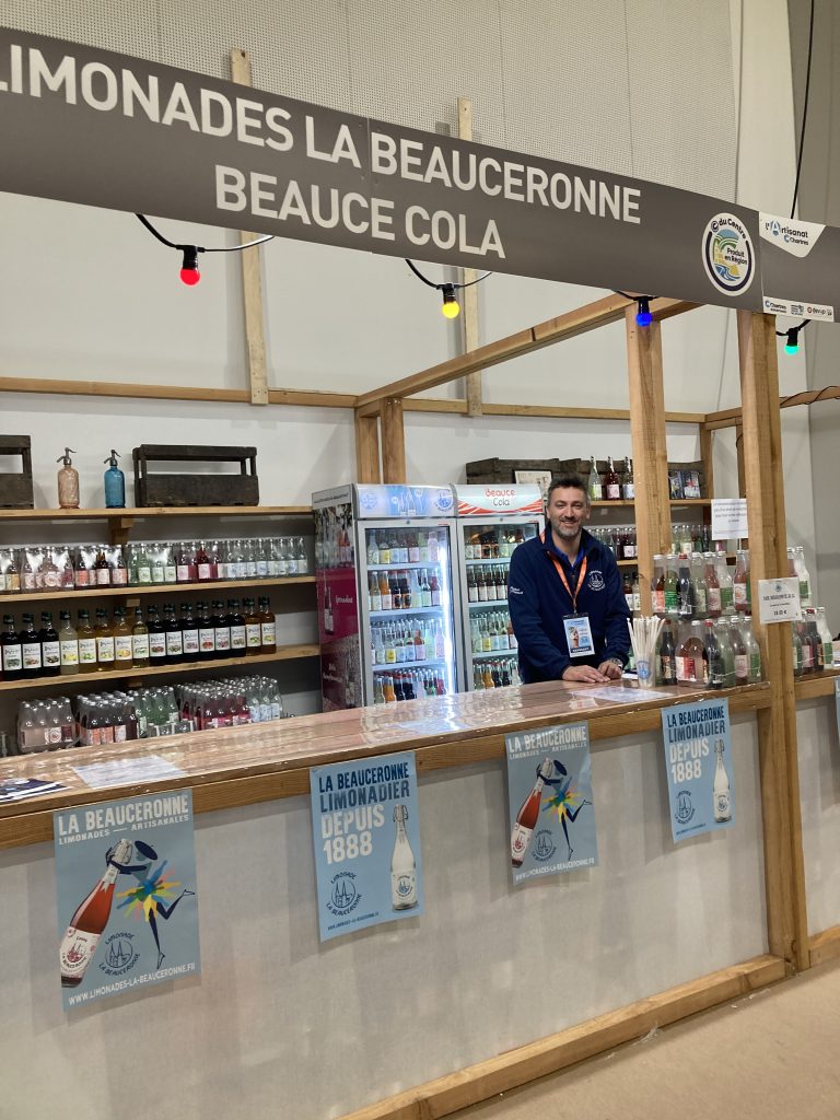 Stand de la Limonade la Beauceronne et Beauce Cola de Maison savouré. Devant se trouve un comptoir blanc couvert d'une blanche bois et derrière trois étagères remplies de limonades et de Beauce cola.