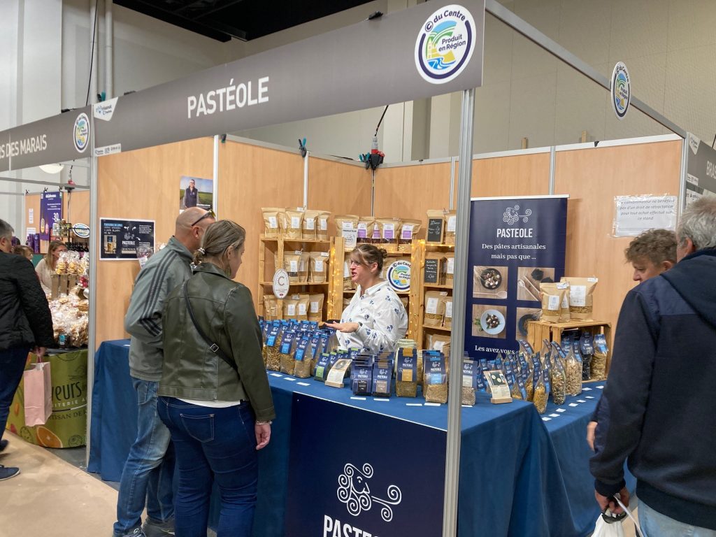 Le stand de Pasteole avec des étagères bois au fond remplies de sachets de pâtes, une kakémono sur la droite et devant une table recouverte d'une nappe bleue sur laquelle sont disposés des paquets de pâtes.