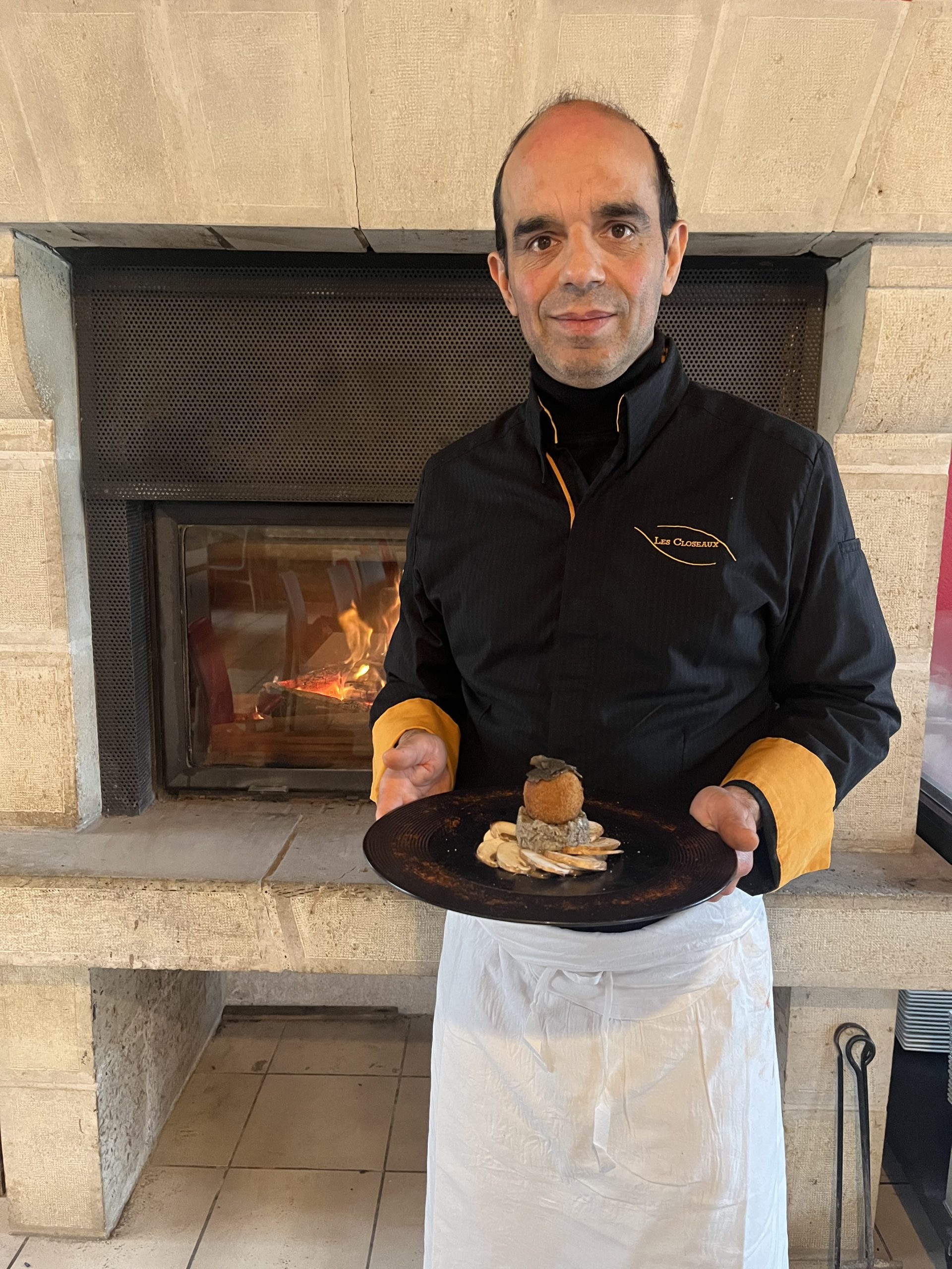 Christophe Lunais, chef des Closeaux, tenant une assiette contenant une entrée festive à base de truffe noire, devant un feu de cheminée.